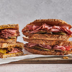 Stack of Venison Pastrami sandwiches with deli meats and pickles on a wooden board against a gray background